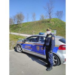 zdjęcie przedstawia strażnika miejskiego w masce ochronnej obok służbowego samochodu